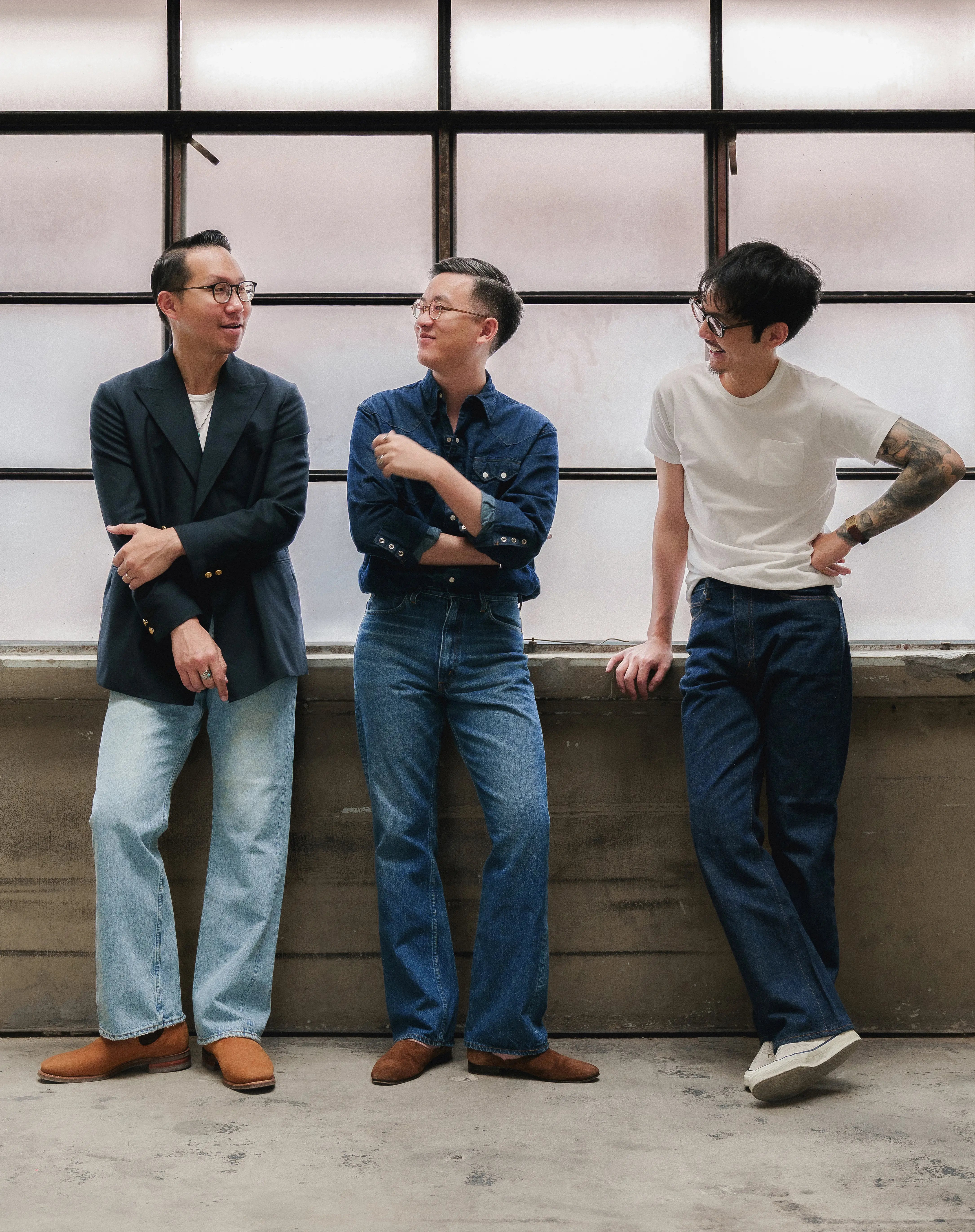 Three men standing and chatting casually indoors by a large window, each wearing different styles of jeans and casual tops — a blazer, denim shirt, and white T-shirt.