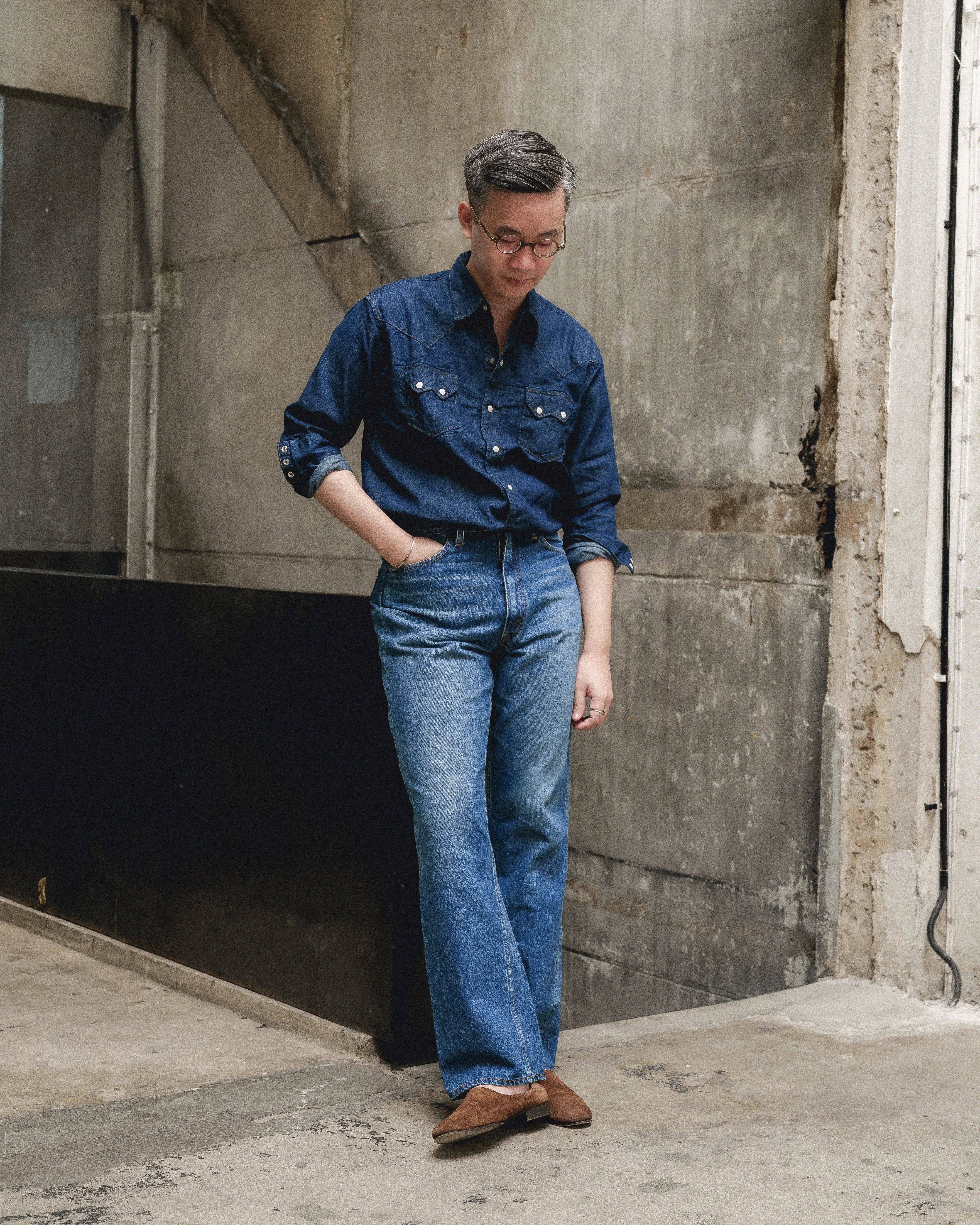 Man wearing Orslow x Clothierandsons 202 Shoe Cut Jeans in 2 Year Wash paired with a dark denim shirt and brown suede shoes, standing against a concrete industrial wall