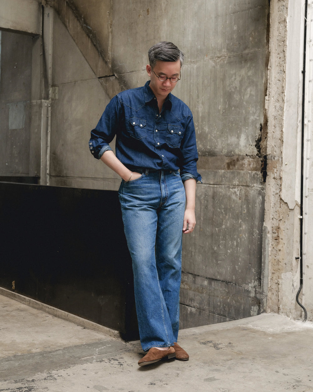 Man wearing Orslow x Clothierandsons 202 Shoe Cut Jeans in 2 Year Wash paired with a dark denim shirt and brown suede shoes, standing against a concrete industrial wall